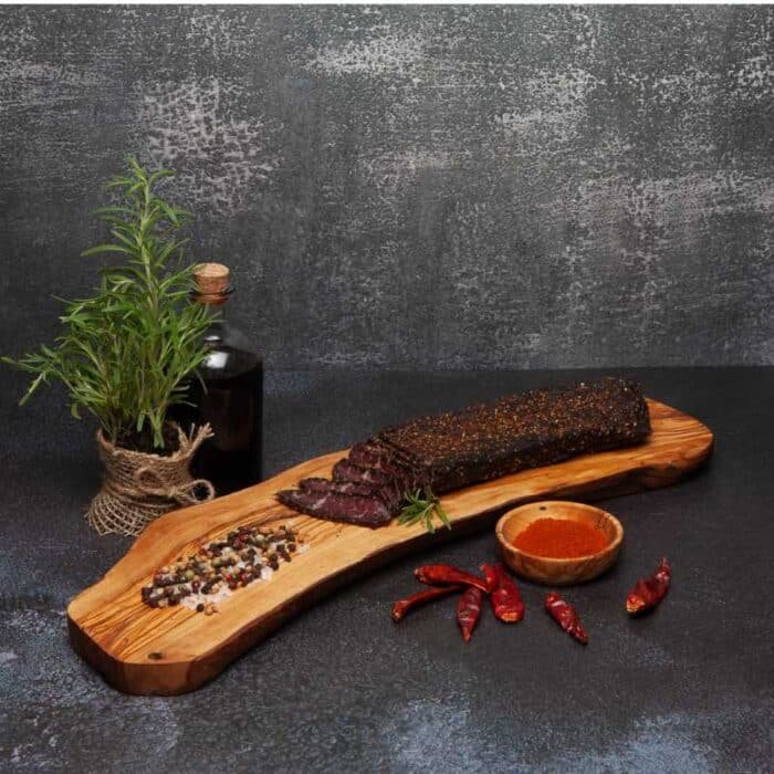 A piece of biltong slab with a few slices on a wooden board next to spices and herbs